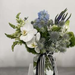 Low glass vase arrangement with white orchids, blue flowers, and green accents tied with a striped ribbon.