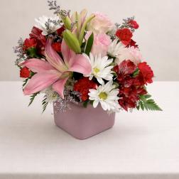 Pink lilies, roses, and daisies arranged in a pink square vase