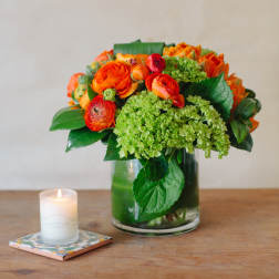 Orange and green floral arrangement in a glass vase beside a lit candle