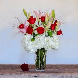 Tall vase arrangement of red roses, pink lilies, and white hydrangeas with one loose rose beside it.