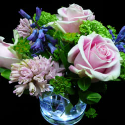 Short arrangement of pink roses, hyacinths, and green blooms in a clear glass vase on a black background