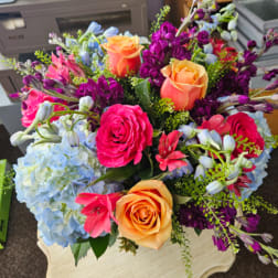 Bright bouquet with orange and pink roses, blue hydrangeas, and purple flowers on a small pedestal table