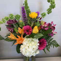 Tall mixed bouquet with white hydrangea, orange lilies, pink gerbera, yellow blooms and green Bells of Ireland in a clear vase