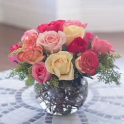 Round bouquet of red, pink, peach, and cream roses in a clear glass bowl vase