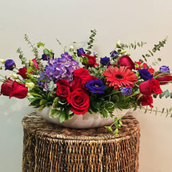 Mixed bouquet of red roses, purple blooms, and a pink gerbera daisy in a white bowl