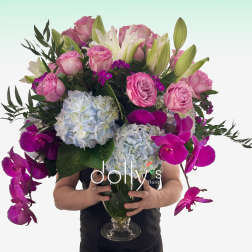 Tall arrangement of pink roses, white lilies, blue hydrangeas, and fuchsia orchids in a clear glass vase