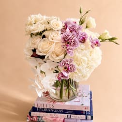 White roses, hydrangeas, and orchids arranged in a glass vase on stacked books