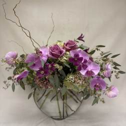 Low arrangement of mauve roses, green hydrangeas, and purple orchids in a round glass vase with tall curly branches