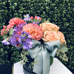 Bouquet of peach roses, purple blooms, and coral dahlias in a black hat box with a blue ribbon.
