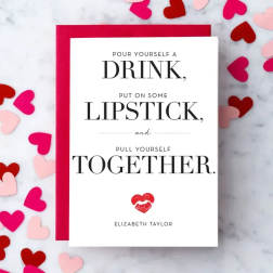 White greeting card with bold black text and red lipstick print, on red envelope with scattered paper hearts.