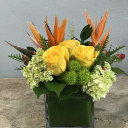 Yellow roses and green hydrangeas in a square glass vase with orange tropical accents