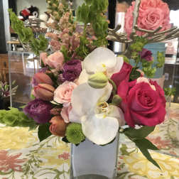 Mixed bouquet of pink roses, white orchids, and purple blooms in a square vase