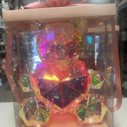 A faceted iridescent teddy bear in a clear gift box with a pink ribbon