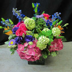 Colorful arrangement of pink hydrangeas, roses, green blooms and mixed purple and blue flowers in a glass cube vase