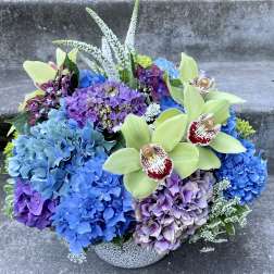 Blue and purple hydrangea arrangement with pale green orchids in a patterned bowl