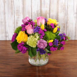 Low arrangement of yellow and pink roses, purple blooms, and green accents in a clear glass vase