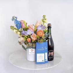 Mixed pastel bouquet in a white vase with chocolate boxes and a bottle of sparkling wine