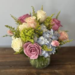 Round arrangement of pink and white roses with blue hydrangeas in a clear glass vase