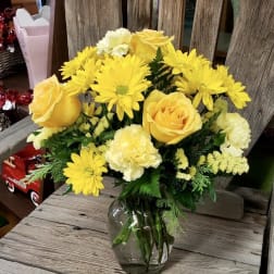 Yellow roses, daisies, and carnations arranged in a clear glass vase on a rustic wooden surface