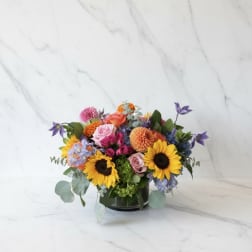 Mixed bouquet with sunflowers, roses, and purple blooms in a low vase
