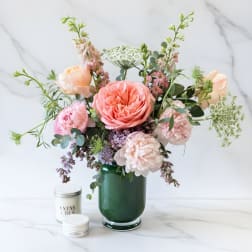 Pastel pink and peach flower arrangement in a dark green glass vase with two small white candle tins.