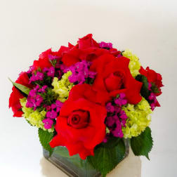 Red roses with pink and yellow filler flowers in a square glass vase