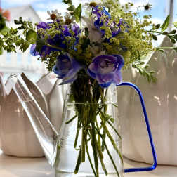 Purple and white flowers arranged in a clear glass watering can vase