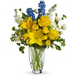 Tall glass vase with yellow lilies, yellow and white roses, and blue delphinium.
