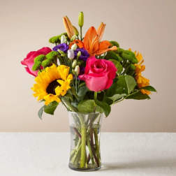 Mixed bouquet of roses, lilies, sunflowers, and purple blooms in a glass vase