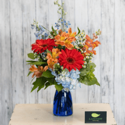 Mixed bouquet of red gerbera daisies, orange blooms, and blue hydrangeas in a blue glass vase