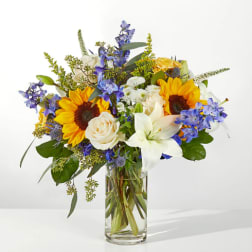 Bouquet of sunflowers, white lilies, and blue flowers in a clear glass vase
