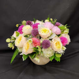 Low floral arrangement of pale roses, pink blooms, and purple accents in a round vase