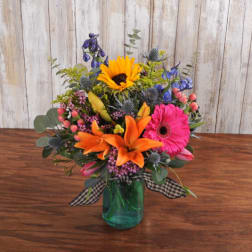 Colorful bouquet with orange lilies, sunflower, and pink gerbera in a green glass jar vase.