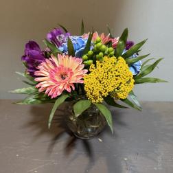 Mixed bouquet of pink gerbera daisies, purple alstroemeria, and yellow filler in a glass vase