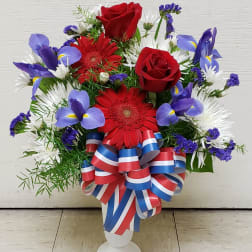 Red roses and mixed daisies in a white vase with a red, white, and blue ribbon