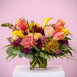 Colorful arrangement of yellow calla lilies, roses, tulips, and protea in a clear glass cube vase