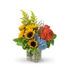 Sunflowers, roses, and blue hydrangeas in a clear square vase