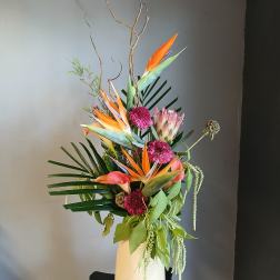 Tall tropical arrangement with orange bird-of-paradise flowers in a white vase.