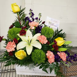 Mixed floral arrangement with yellow roses, white lilies, and pink carnations in a white container
