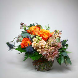 Orange roses arranged in a glass bowl with blue thistle and pale pink blooms