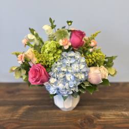 Compact arrangement of pink roses, blue hydrangeas, and mixed blooms in a white vase.