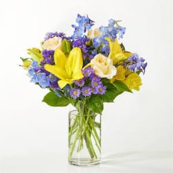 Bouquet of yellow lilies, white roses, and blue-purple flowers in a clear glass vase