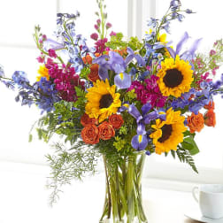 Colorful mixed bouquet in a clear glass vase with sunflowers, roses, and blue flowers