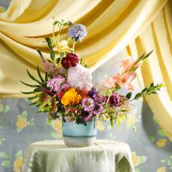 Colorful mixed flower arrangement in a blue ceramic vase on a draped table with yellow fabric backdrop.