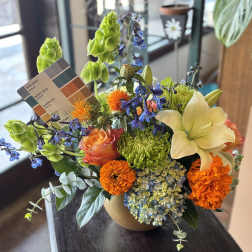 Mixed bouquet in a tan vase with orange, blue, green, and cream flowers.
