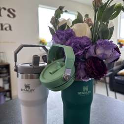 Purple and white flowers arranged in a green Stanley tumbler beside a white tumbler