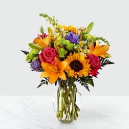 Mixed bouquet of sunflowers, roses, lilies, and purple blooms in a clear glass vase