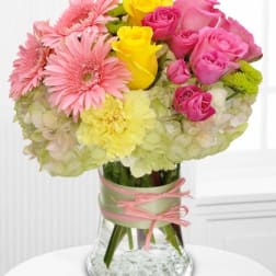 Compact vase arrangement with pink gerbera daisies, pink and yellow roses, and yellow blooms over white hydrangeas