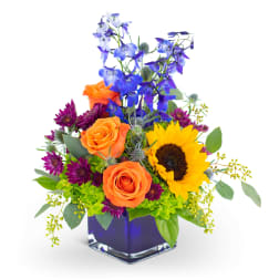 Colorful bouquet with orange roses, a sunflower, and blue flowers in a square vase