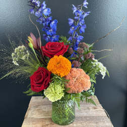 Mixed bouquet with red roses, blue delphinium, and orange blooms in a green glass vase